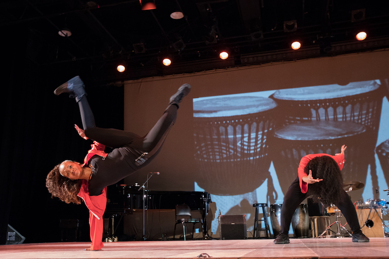 A b-girl lifts her body off the ground with one hand. Her legs are bent and above her head. Another dancer bends her torso over her legs which are in a deep squat.