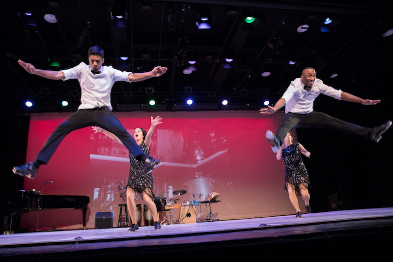 Two dancers splay their legs and leg into the air. Women wearing flapper black bedazzled dresses raise their arms behind them.