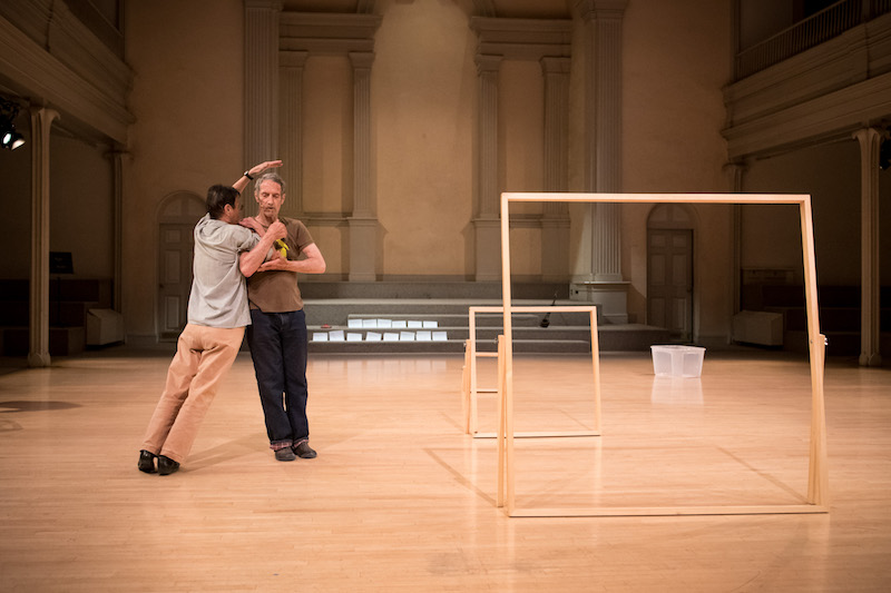 Steve Paxton stands erect as Yvonne Rainer leans against him. To their left, are three wooden picture frames of varying heights.