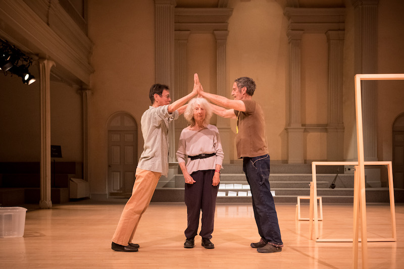 Yvonne Rainer and Steve Paxton press their hands together making a roof top shape. Simone forti stands between them underneath their hands. 