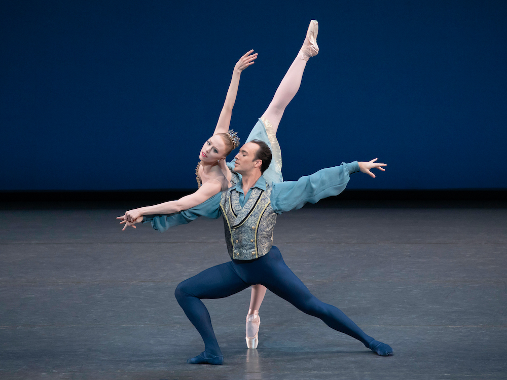 a pale red headed ballerina with a tiara leans into her male partners arm in a long arabesque. We can see she wears a costume of bably blue, her partner a male with black hair, sightly darker skin, and donned in a blue top, brocade vest,and dark blue tights supports her weight with an air of gallantry.