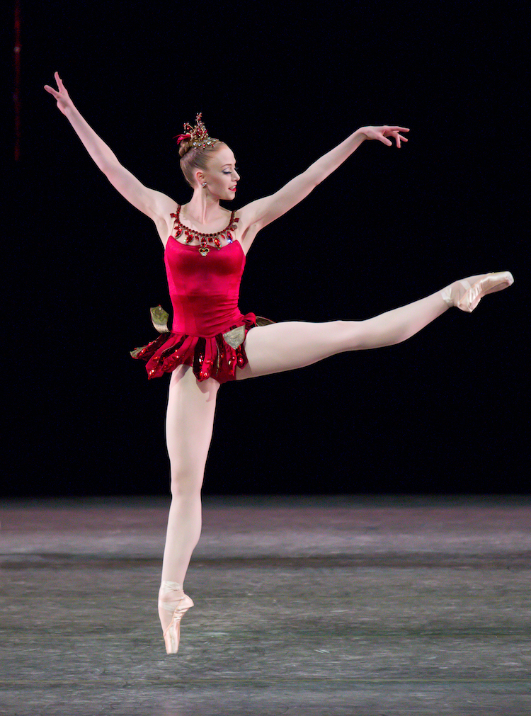 a fair skinned ballerina in a bright red ruby reminiscent tiara and a bejeweled velvet leotard with a whimsical mini skirt attached jumps jazzily in the air one leg extended to the floor, and the other thrust out to her left side.