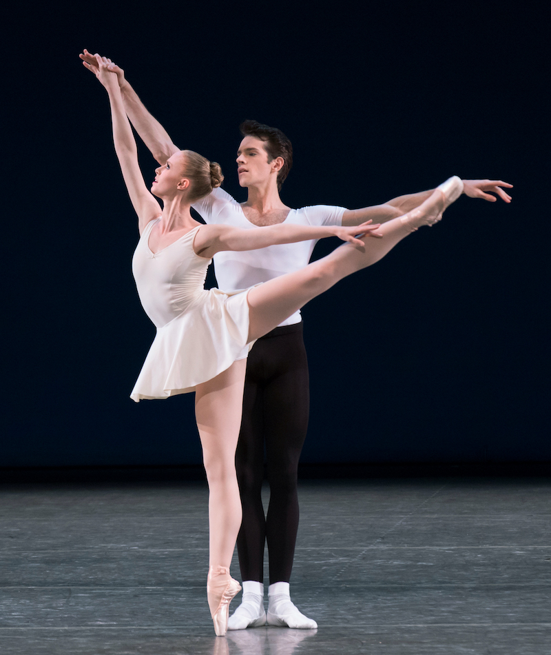 a ballerina in a white tunic gives us a side view of an extra high arabesque, her male partner in a white tshirt black pants and white ballet  shoes stands steadily behind her supporting her with a gentle held hand