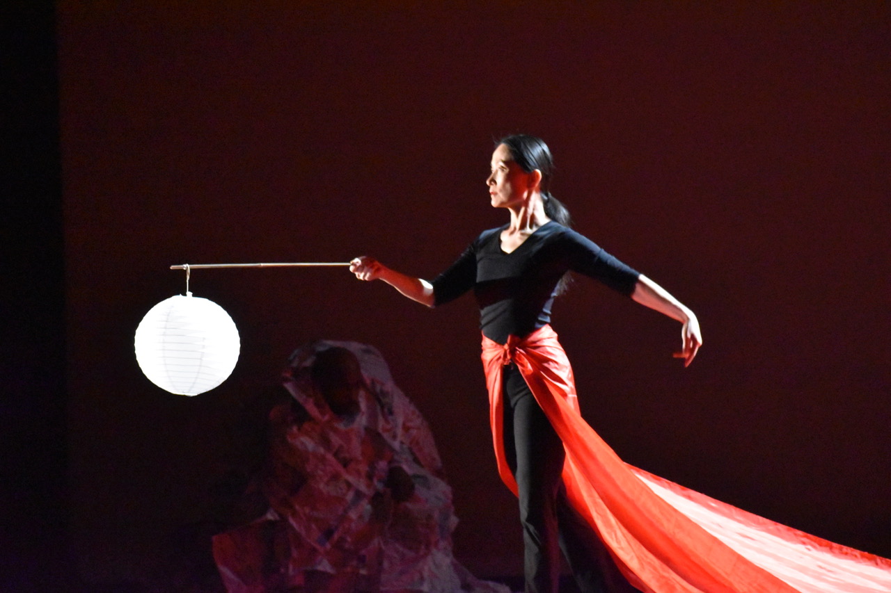 Nai-Ni Chen in performance,an Asian woman with black hair ,in a black leotard with long sleeves and an orange long skirt holds a lantern in front of her as she moves serenly forward
