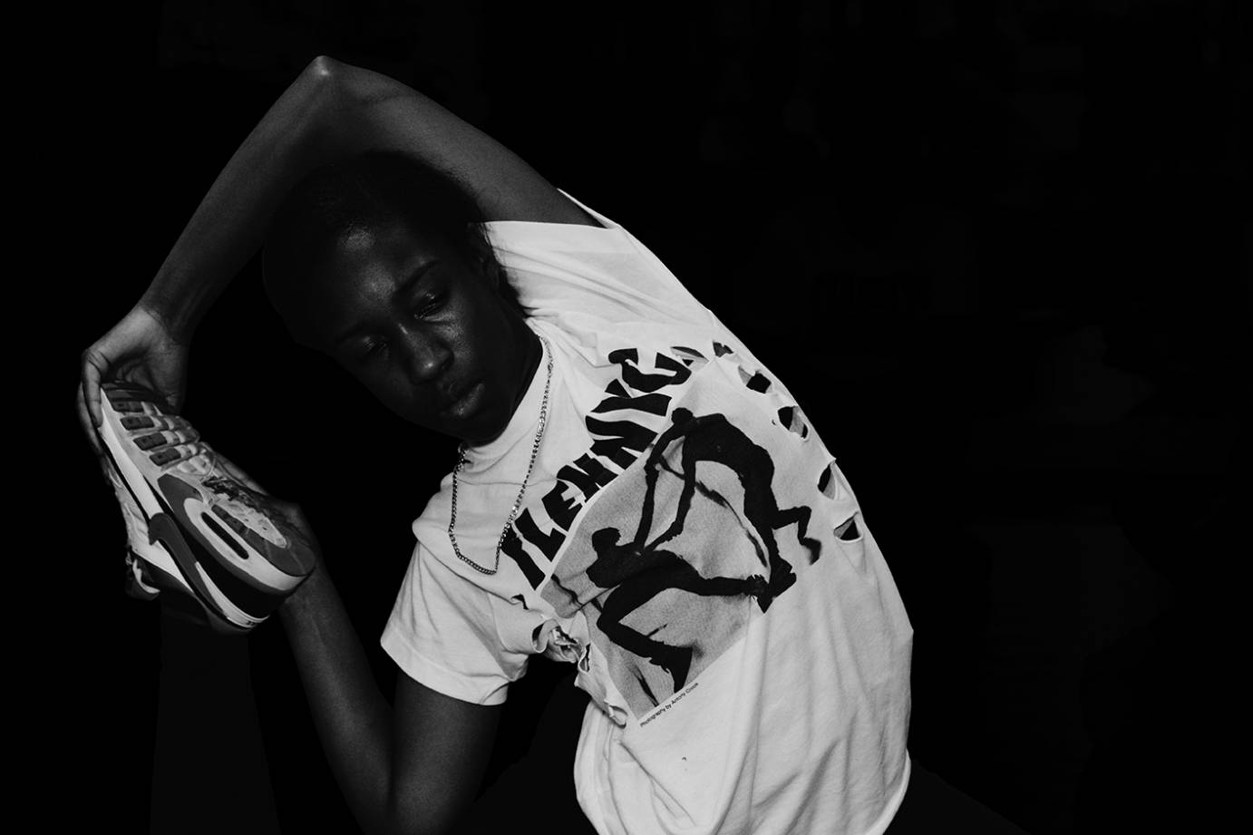 A black and white photograph of a dancer wearing a FlexNYC t-shirt and stretching his obliques while holding a sneaker.