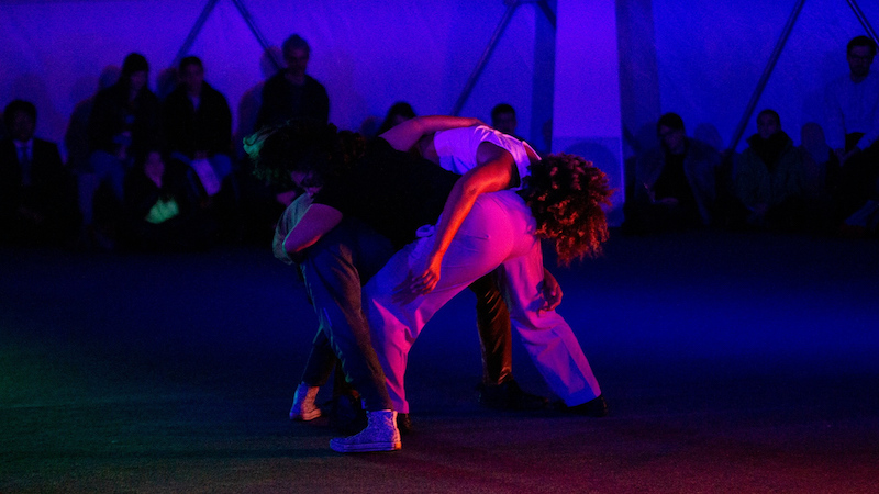 A woman folds over an individual's shoulder as shadowy figures lurk on the sidelines. Everyone is bathed in violet light.