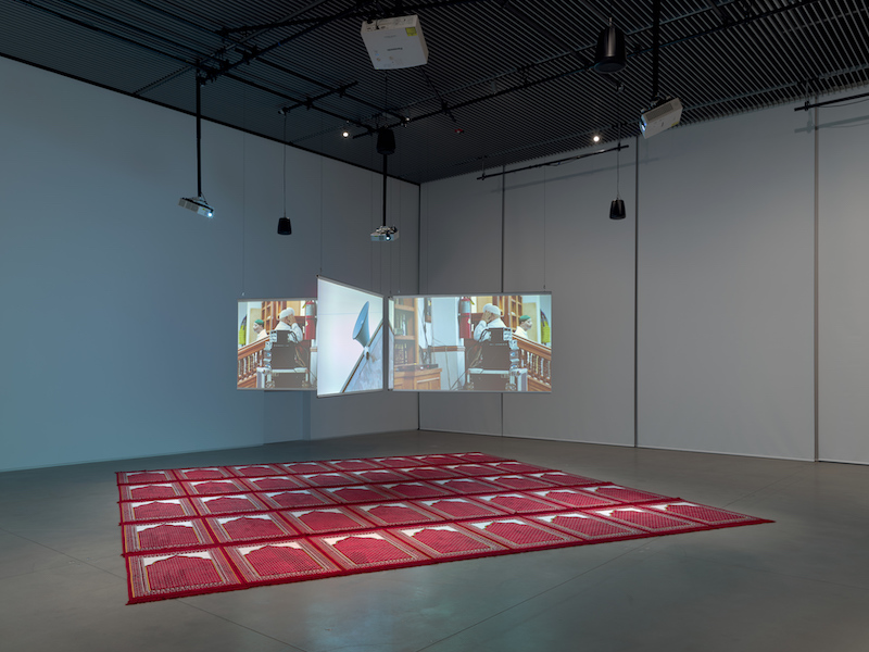 A three-paned video installation hangs from the ceiling. The panes are open like leaves in a book. A red and white carpet-like covering lays underneath. 