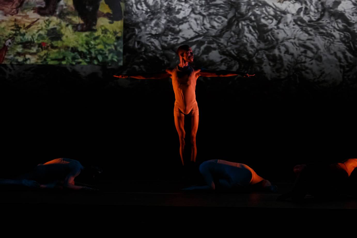 A man drenched in orange light stands before a mysterious forest. The background is dark.