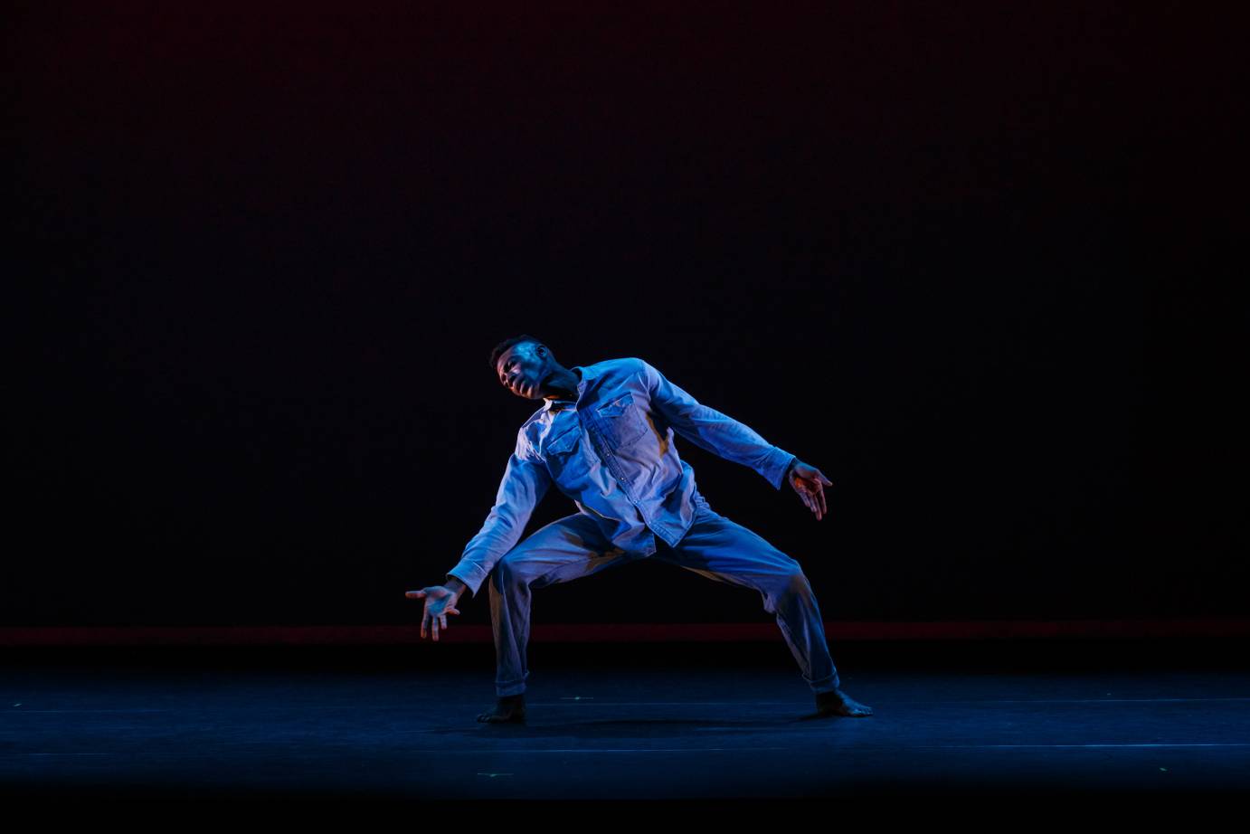 A man, in a deep 2nd position, tilts his body and lunges to the left, his arm in a beseeching gesture.