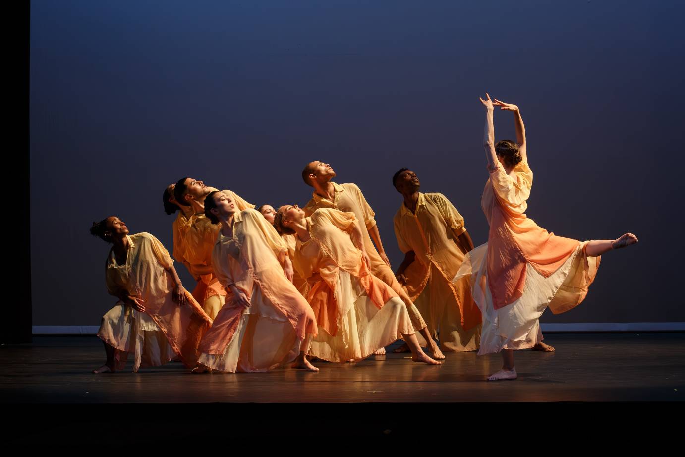 The company, costumed in light orange and cream, leans back as a dancer faces them with arms up and leg behind in arabesque.