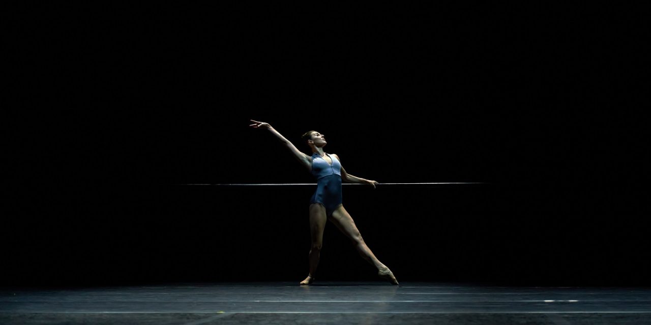 In a blue leotard, Tiler Peck holds on to the barre while extending a leg in tendu devant with one arm lifted to create a diagonal line from finger to toe 