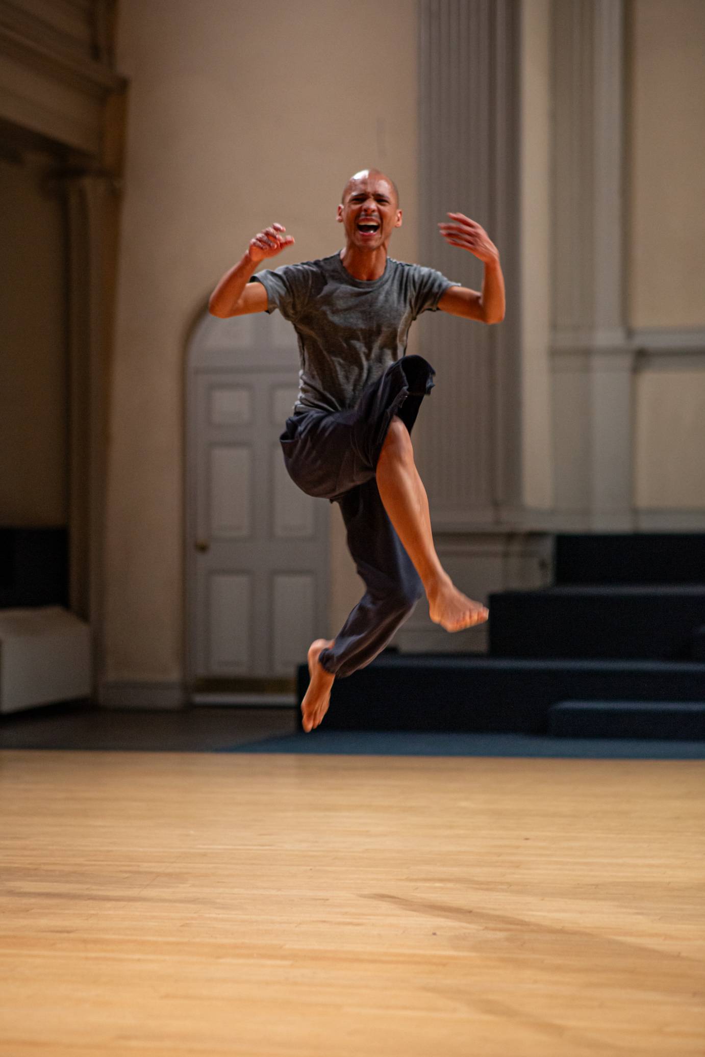 A casually dressed bald headed man grimaces broadly, jumps with one bent leg forward and the other back, elbows bent, hands soft.