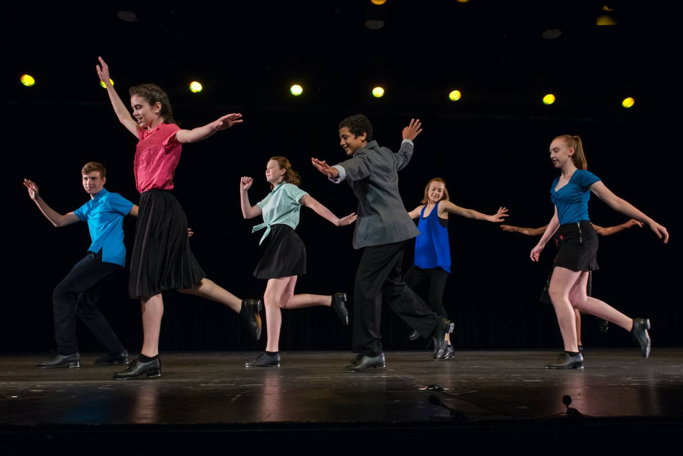 a group of young students joyfully tapping on stage  wearing bright solid color tops and black bottoms