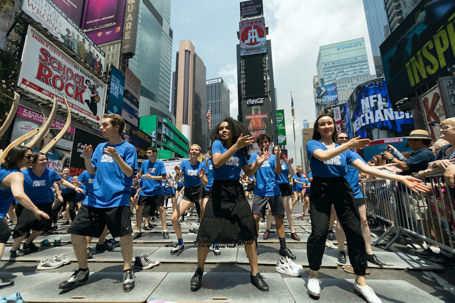 a group of young tappers tap on the streets of Time Square as an appreciative audience looks on 