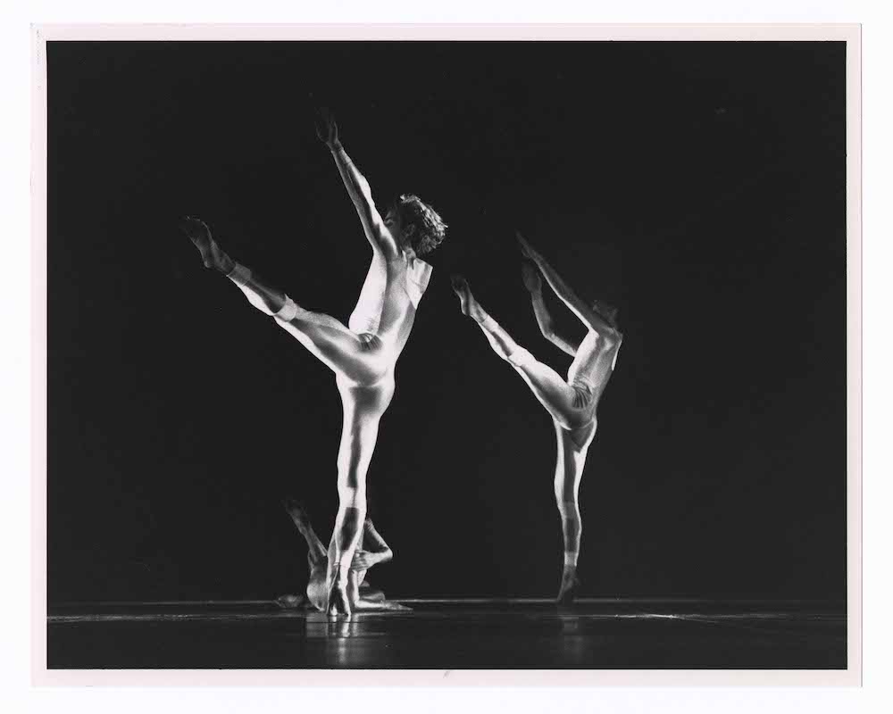 black and white photo, 2 dancers in unitards, facing back, kick their legs high to their sids