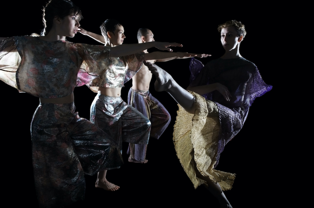 three dancers in glittery costumes extend their arms in a line while another dancer kicks her leg out as if to reach out to their arms.