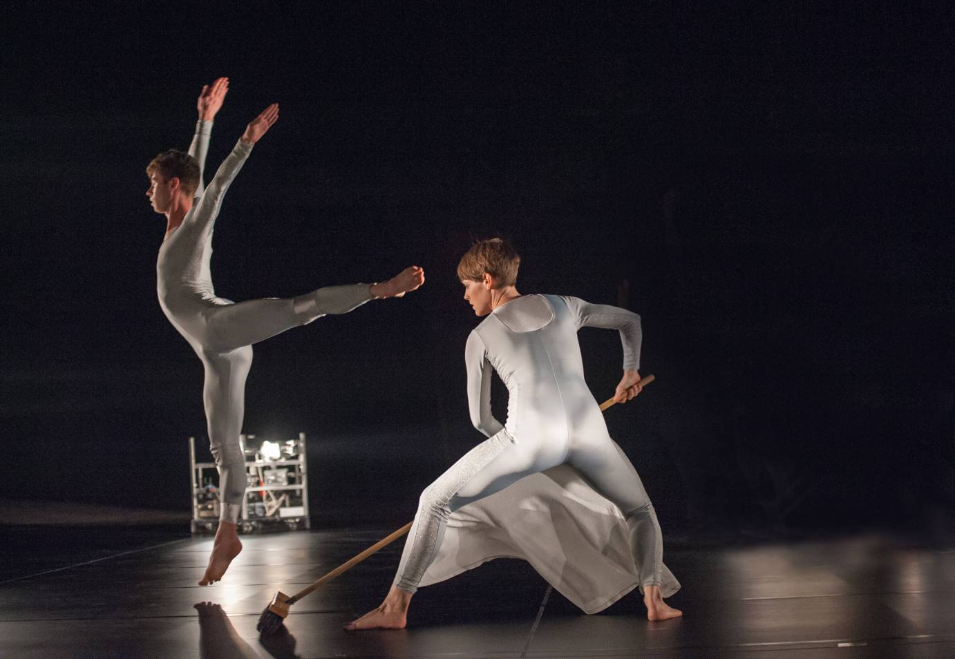 a dancer in silky grey-blue unitard jumps in the air as another dancer sweeps with a long broom