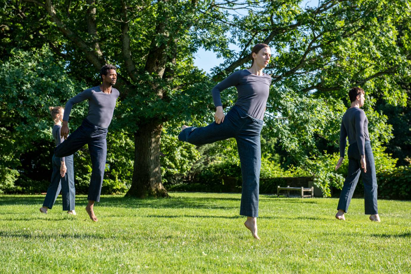 Two dancers in dark pants and top jump in attitude while two other dancers stand in releve in profile