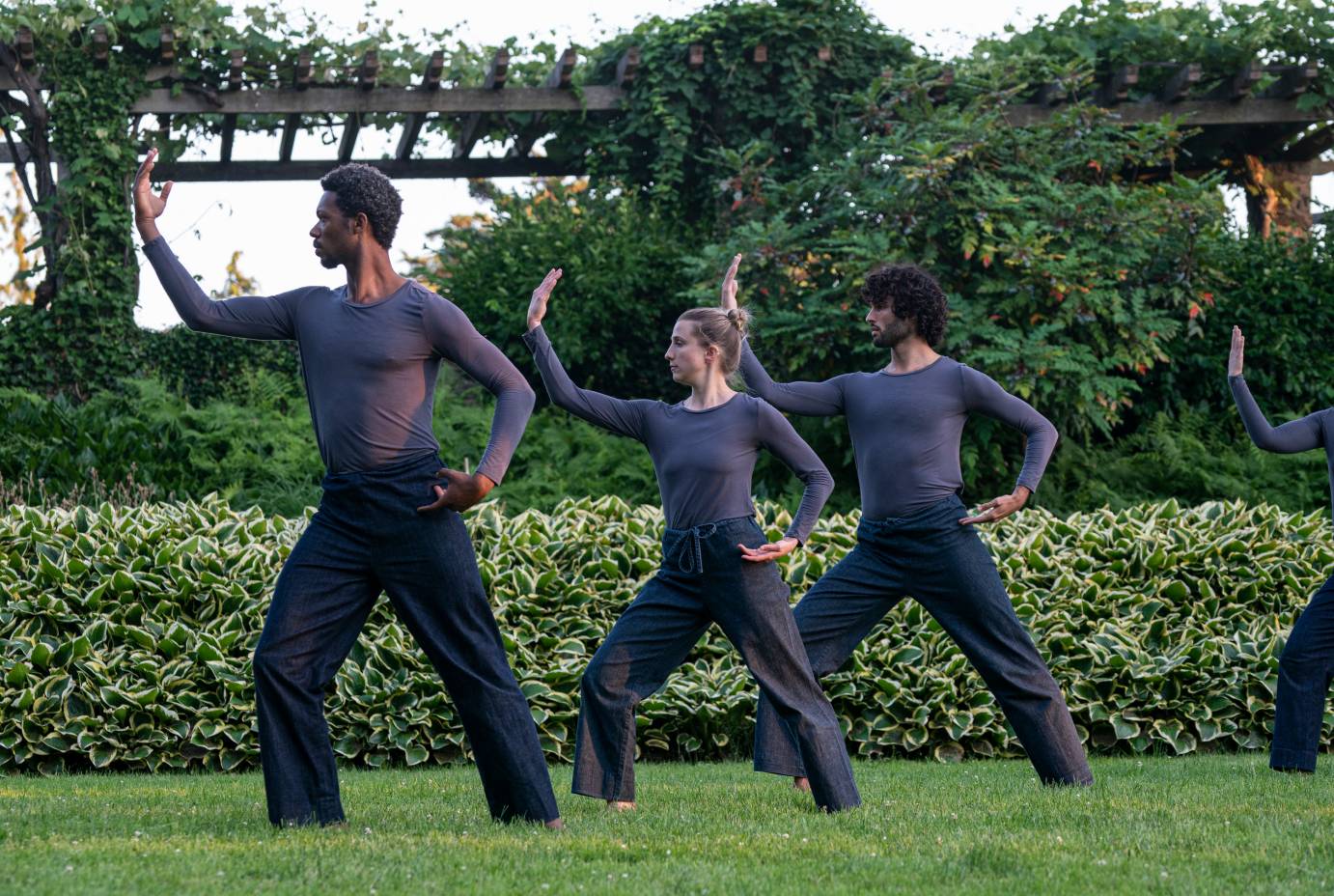In a beautiful outdoor setting of trees and bushes, three dancers stand in profile with one arm rounded upward and another arm rounded at the hip