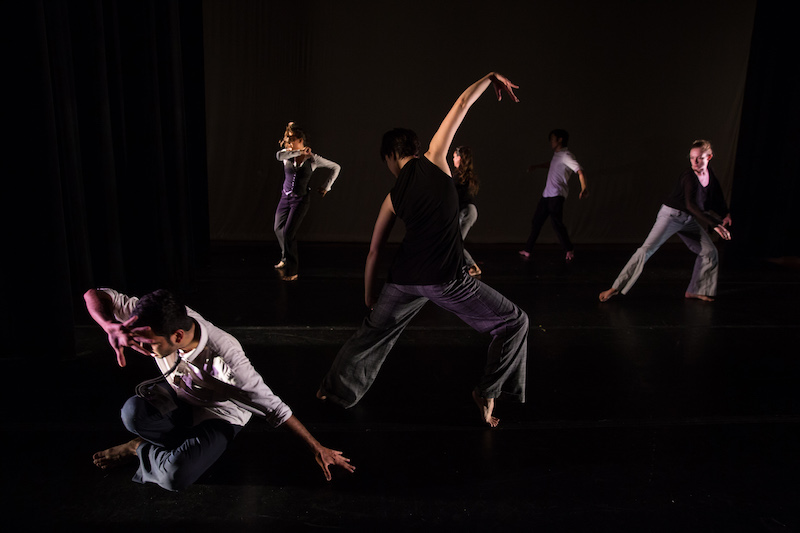 A group of dancers in business-wear strike various poses on a darkly lit stage.