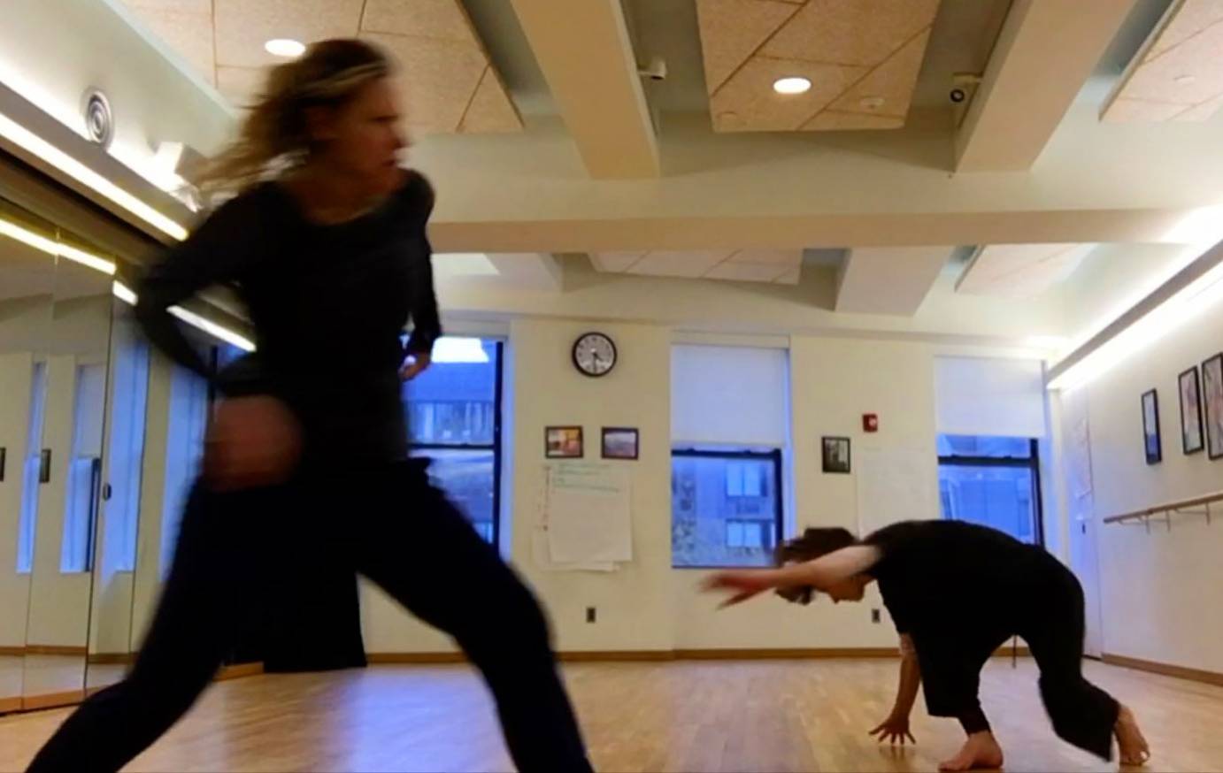 Two women in rehearsal clothes dance in a studio. One is spiral to the ground while the other is in a deep lunge.