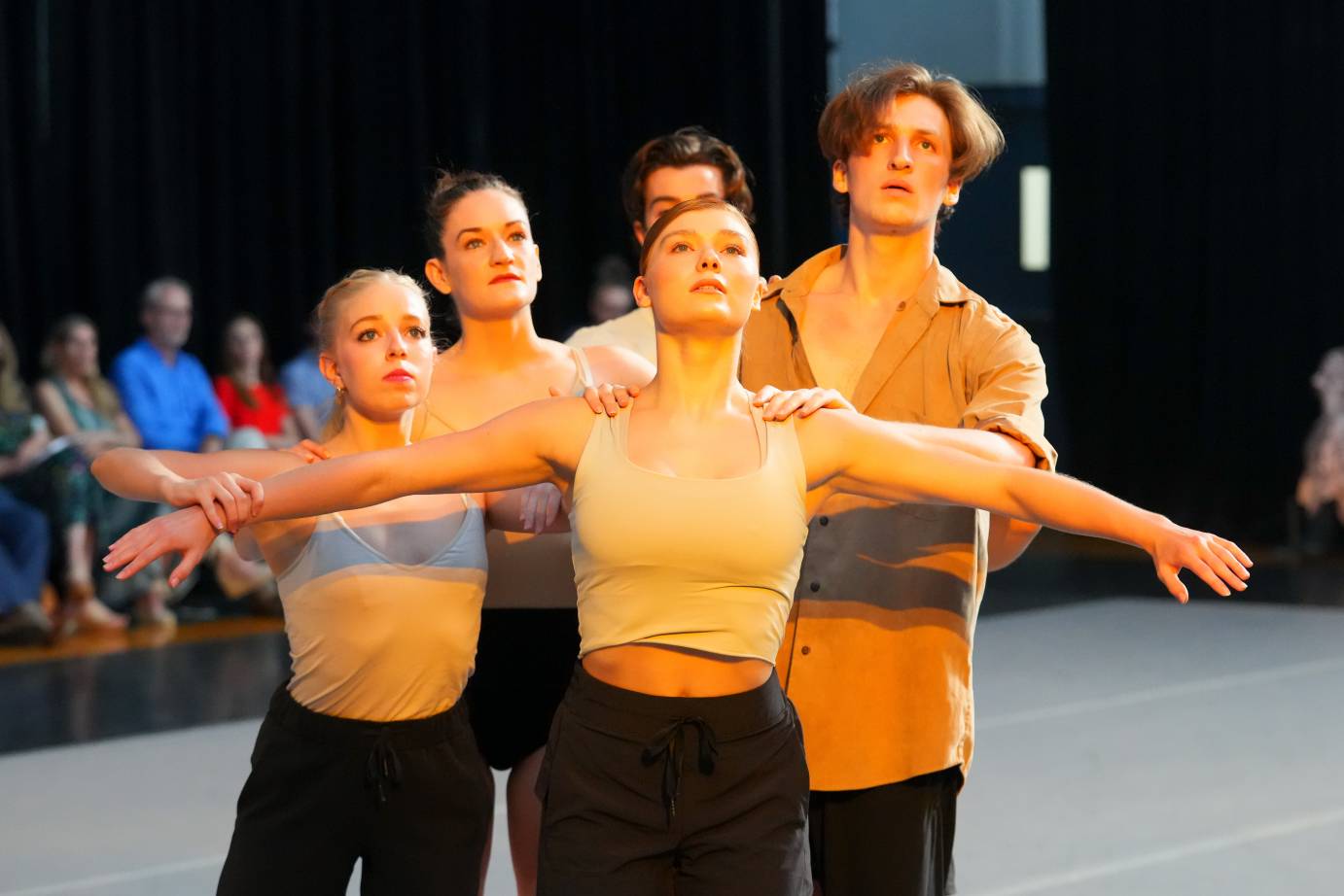  Haley Winegarden heads a group of fair skinned dancers in various styles of gold tops and black bottoms stare out into space with a sense of wonder