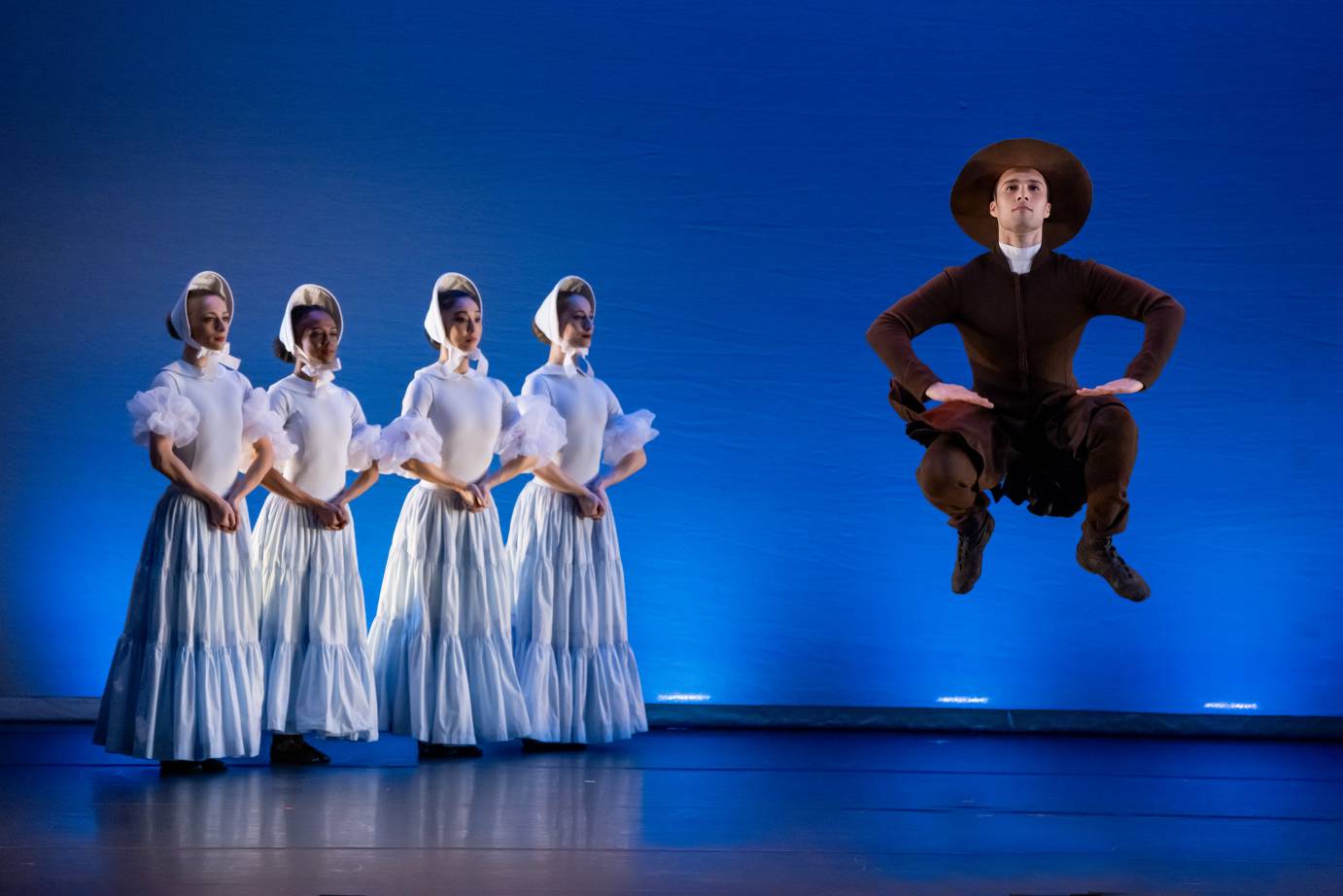 4 female dancers holding hands upstage. To their left a male dancer is jumping with 2 legs bent