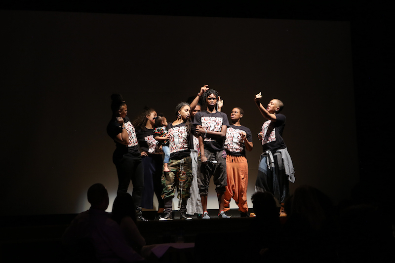 A woman stands with her arms across her chest while a group of dancers stand around her gesturing towards her 