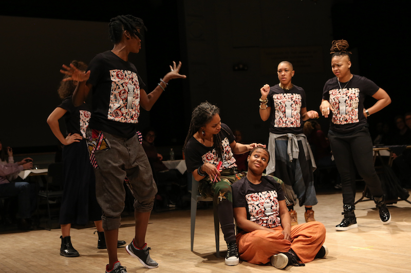 One woman sits in a chair and strokes another woman's scalp. Other performers stand around them. 