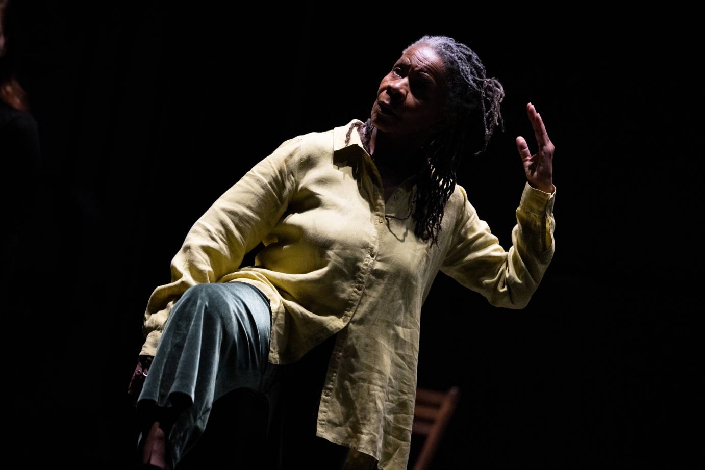 Bebe Miller, a black woman dancer choreographer stands on one leg, tilting her body slightly to stage right, she is wearing a long tan collared shirt with long sleeves and velvet light blue pants... 