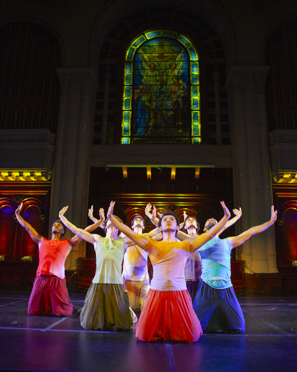 Six men dancers in bright costumes with uplifted arms and on their knees in front of a Tiffany glass window depicting Christ healing children