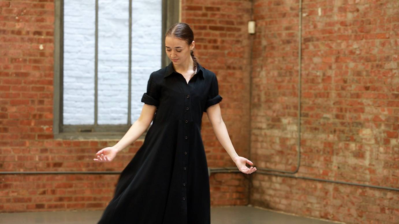 dressed in a black dress, Valerie Kosnevich, in a moment from her choreography, looks downward at her open palms as if in meditation