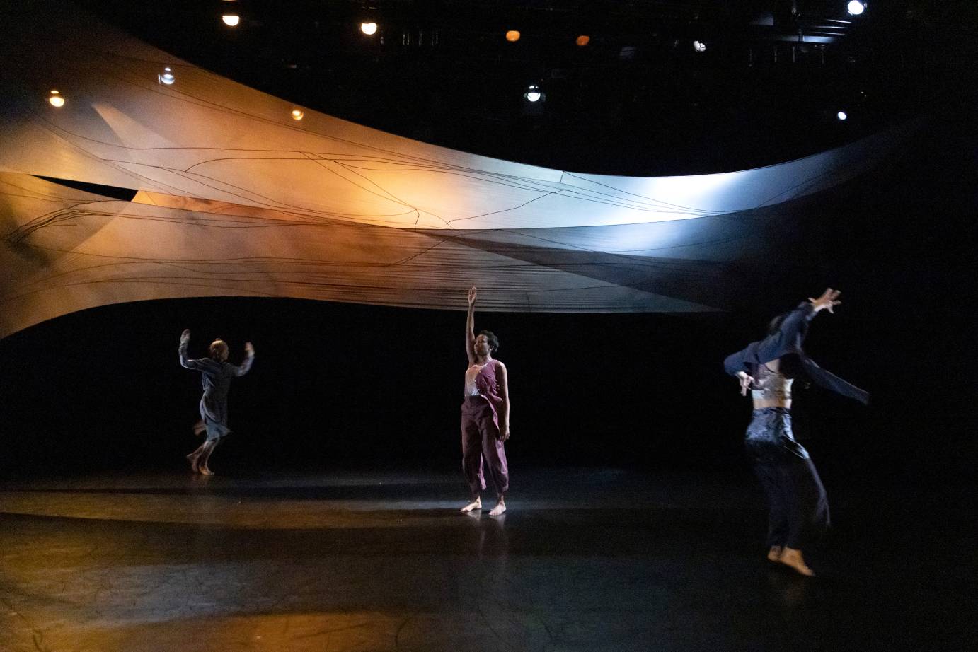 Three performers equally separated and in a line beneath the luminous set stretched above them, hop, stand, and skip with arms reaching up and out.