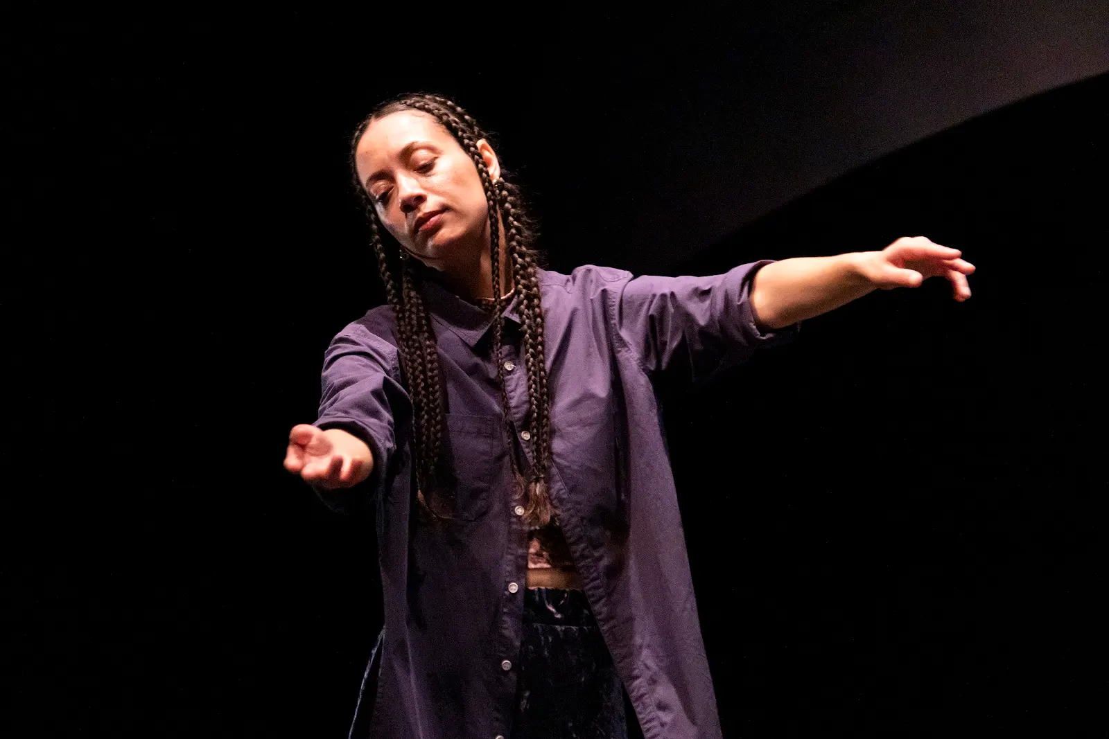 A woman with long braids and wearing a purple shirt, tilts her head toward her shoulder, eyes downcast and face relaxed, arms stretched toward the audience