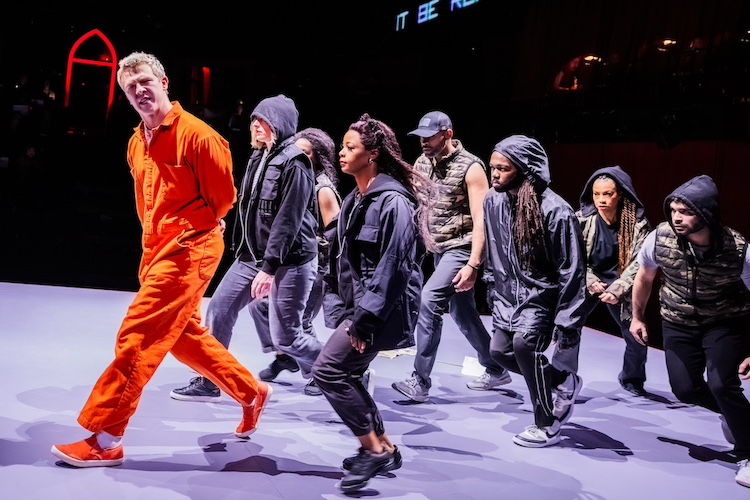 a white-colored man in an orange prison jump suit with matching shoes, leads a group of people in darker clothes, many in hoodies or caps, in a procession. He seems in the middle of saying something important as the others are quiet.