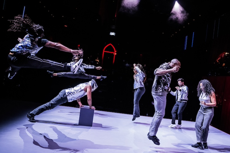 a vibrant scenee of dancers engaged in activity on the stage. two are facing eachother in the air while their legs extend out to each side of them. Below them is another dancer in the midst of an elevated push up on a block. Two more pairs of dancers move together, some slightly in the air the others on their feet on the floor. The dancers dazzle in black pants and silver, mylar like tops. 