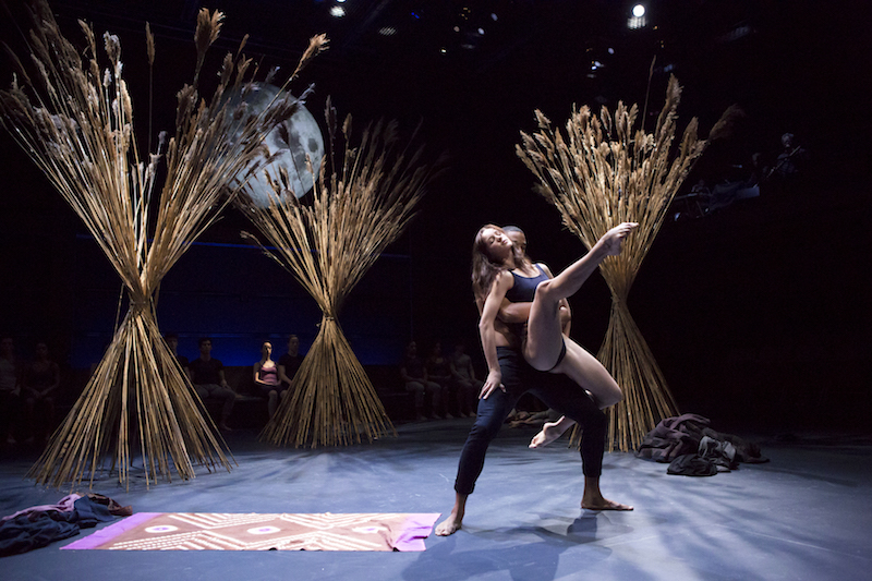 Two dancers in the foreground. A man clasps his hands around his partner's waist. She extends her leg in a high attitude.