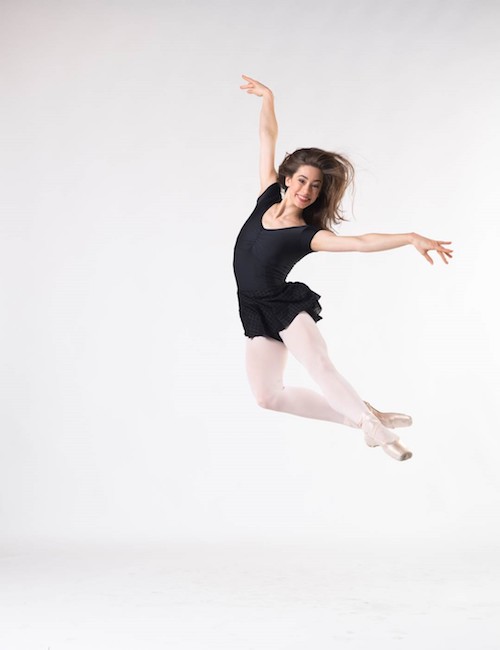 A woman in a black leotard and skirt leaps into the air. Her hair is loose.