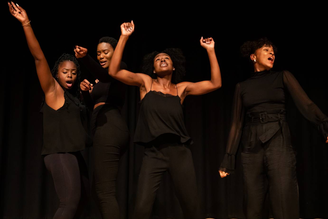 Four Black women dressed in black costumes - over the shoulder, sleeveless, to the chin. Center woman both arms upraised with bent elbows facing the audience, woman on left right arm extended on diagonal, woman on right with head turned over left shoulder, fourth woman head tucked
