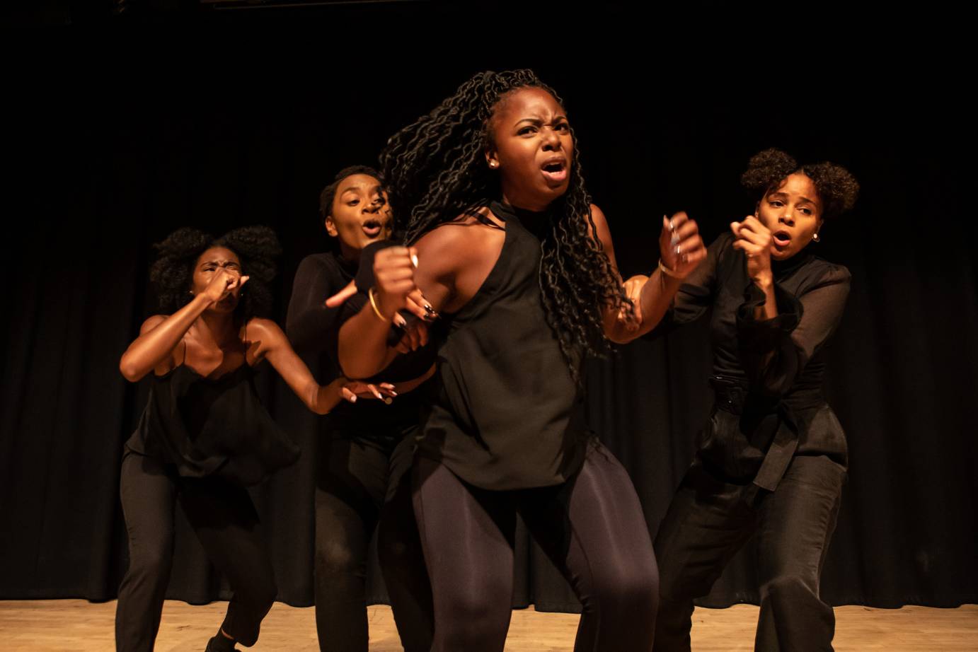 Four Black women - one woman elbows tucked to sides forward of the others who look on with with hands in front of their faces.