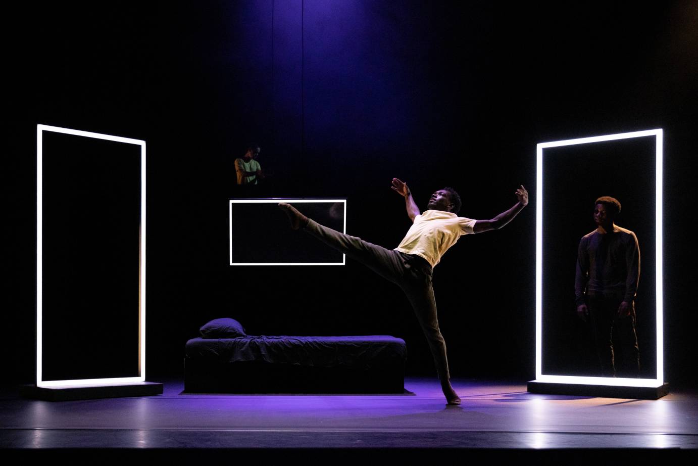 Felisberto, arms horizontal, leg extended forward in bedroom surrounded by three set pieces rimmed in white light