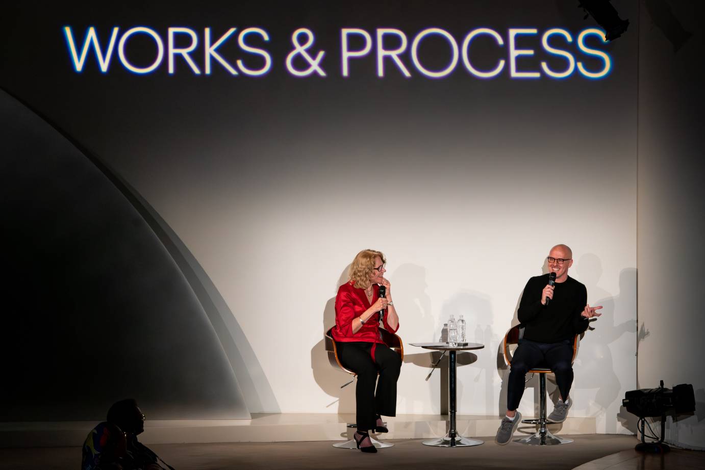 A seated woman and man holding microphones converse. The man is smiling.