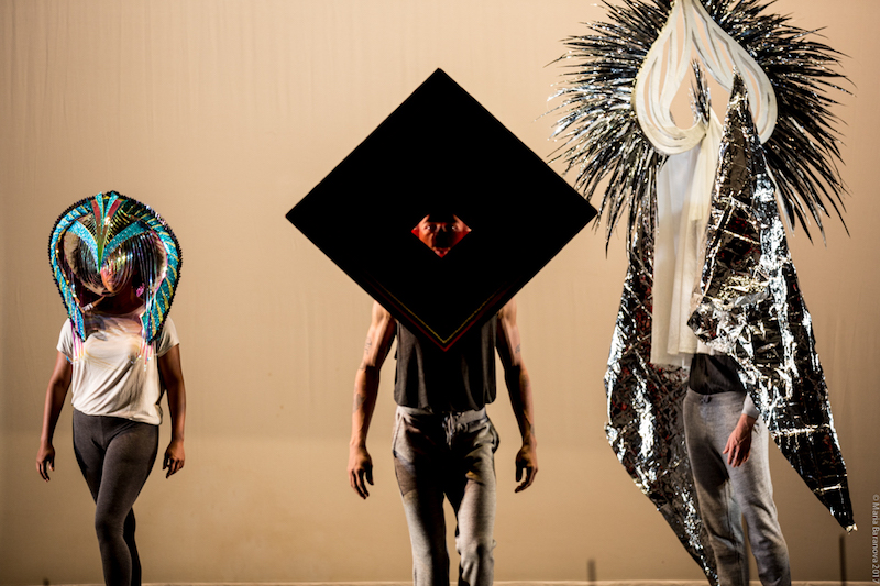 Three dancers face the audience wearing elaborate masks. Some have fringy paper, are shiny, are geometric and one is very tall.