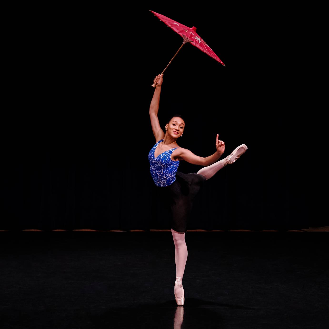 A woman poses in attitude with one hand holding a red parasol while the index finger of her other hand points upward