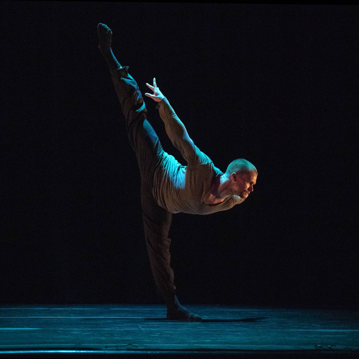 a light skinned man in dark sweat pants and a loose lighter colored long sleeve shirt dives into a penchee arabesque. We have a profile view