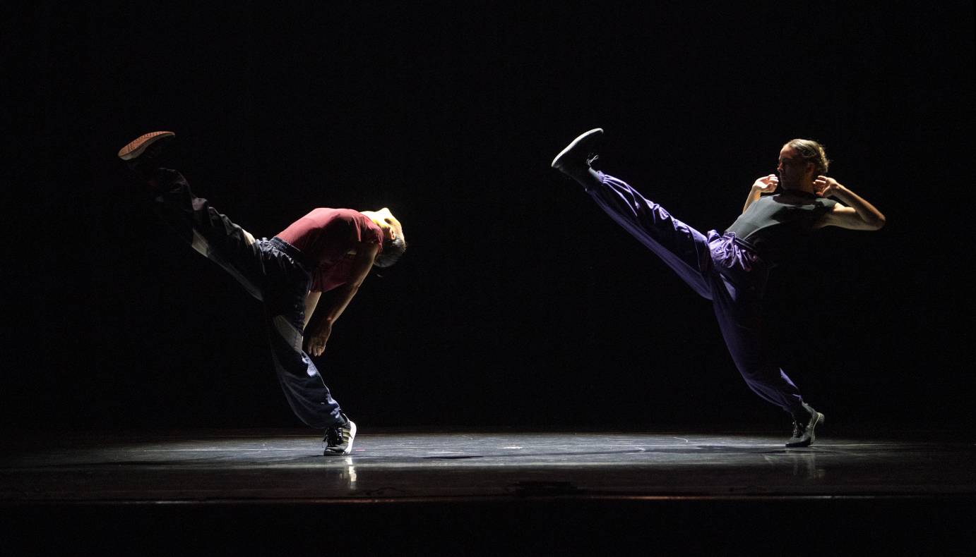two dancers in sweats kick their legs high into the air leaning back away from the leg...
