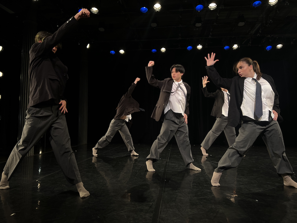 a company of young dancers male and female stand together in unison, legs in a wide stance, one hand on their hip the other in an upward motion as if to say