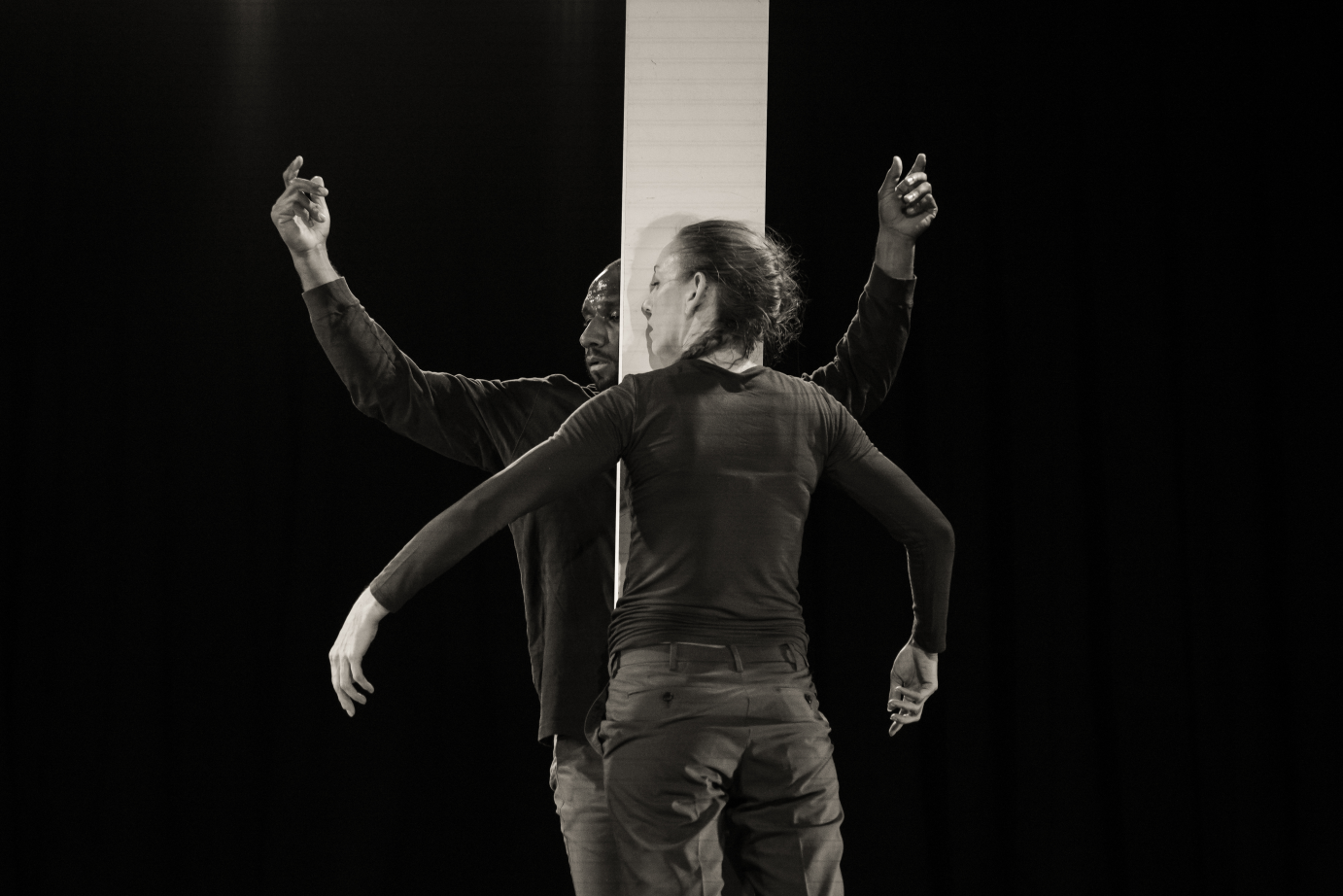 A black and white photo shows Nik Owens and Amber Sloan with a board between them. Standing, they press themselves against the board. Sloan's arms curve downward while Owens' curve upward.