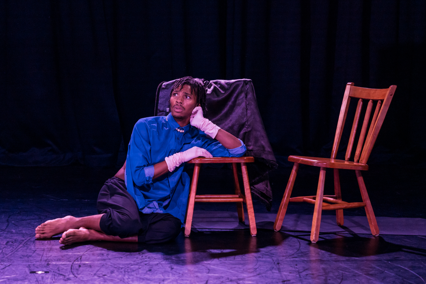 In a blue blouse, white gloves, and pearls, Sy Lu rests an elbow on a wooden stool. They have their chin in their hand.