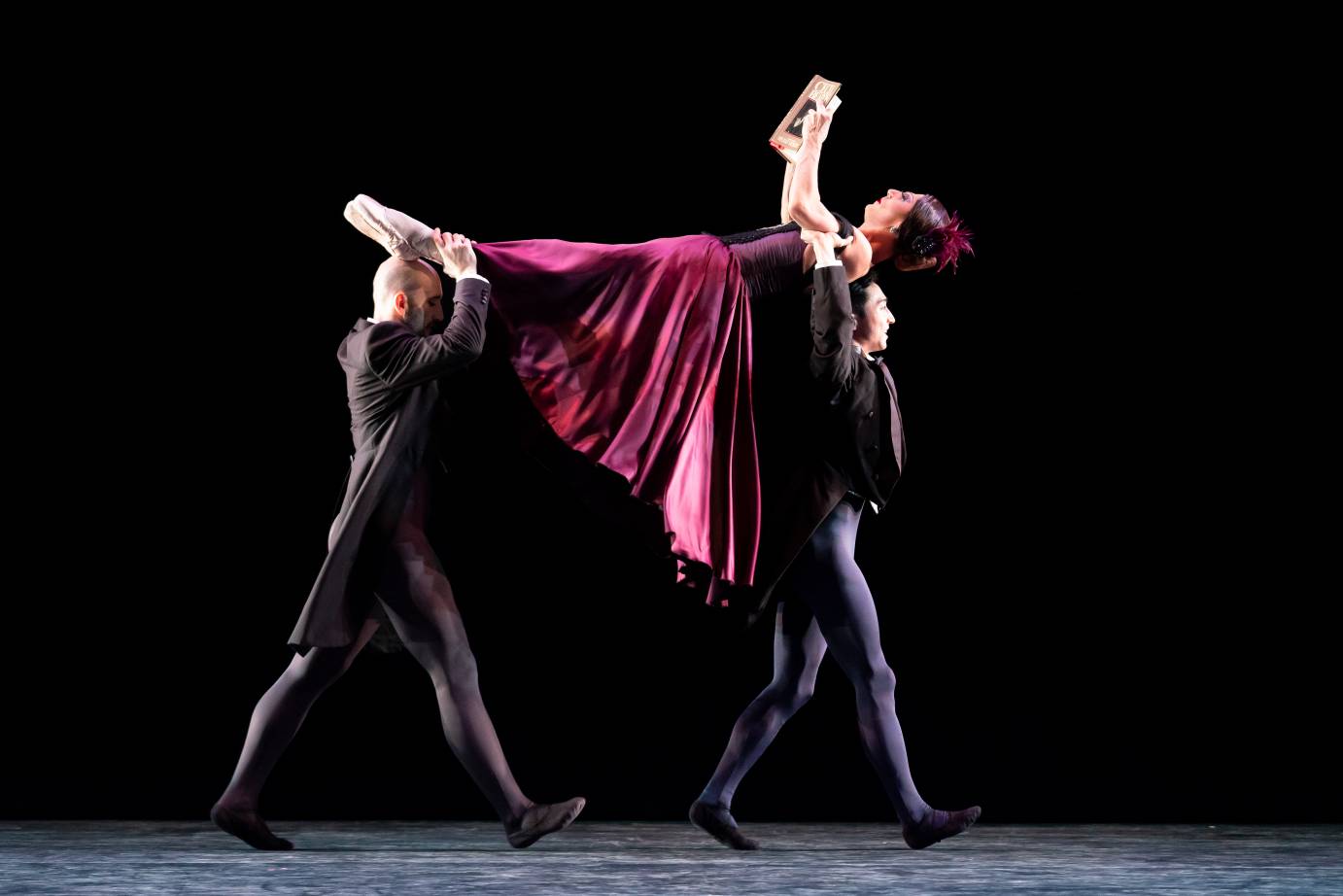 Two tuxedo-clad men carry a dancer in a scarlet dress over their head. The dancer is reading a book.